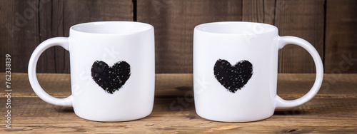 Two white coffee mugs with black heart-shaped prints placed on a wooden table against a wooden background.