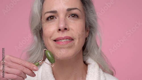 A woman with gray hair is using a jade roller on her face, smoothing her skin