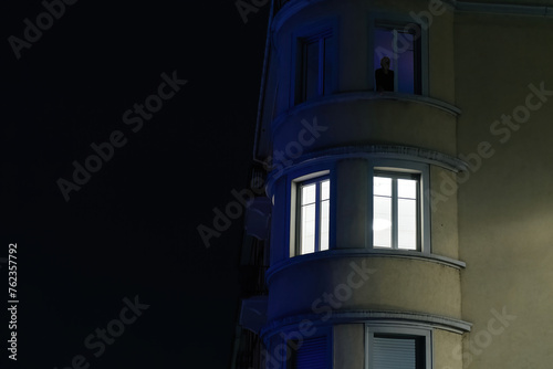 A lone unrecognizable figure looks out from a brightly lit window at night smoking a cigarete