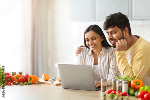 Happy loving young indian spouses using laptop while cooking photo