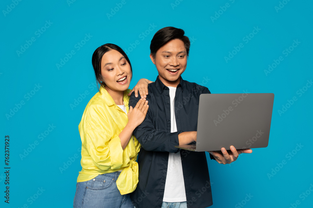 Exuberant Asian couple sharing a laptop screen, the woman leaning on the man's shoulder with a joyful smile
