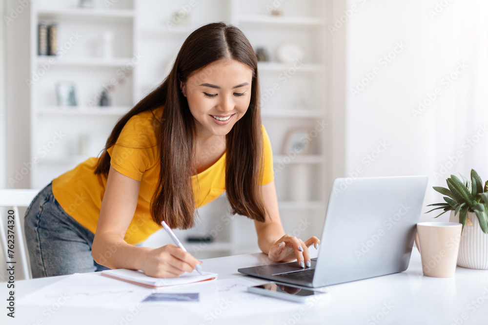 Smiling asian woman taking notes while working on her laptop at home Smiling asian woman taking notes while working on her laptop at home