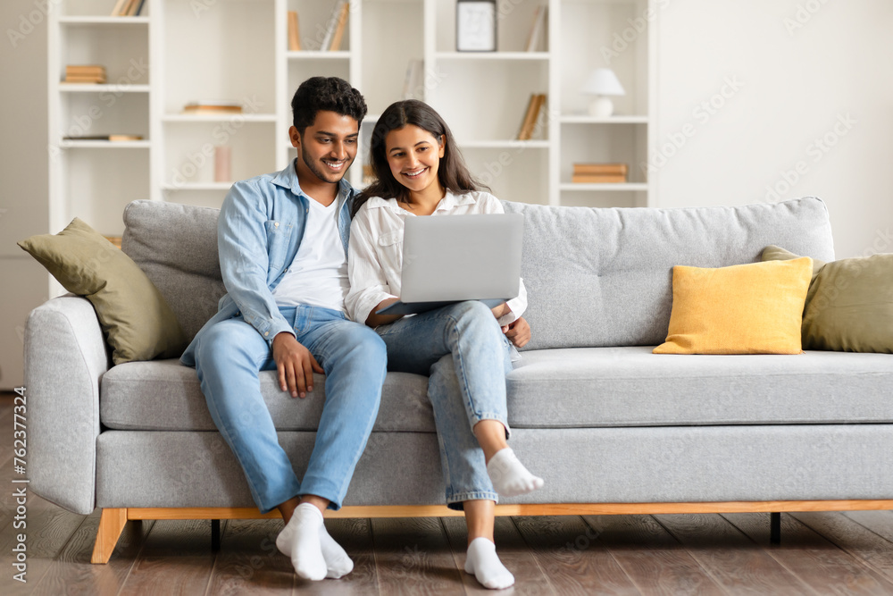 Relaxed indian spouses resting on couch with laptop smiling Relaxed indian spouses resting on couch with laptop smiling