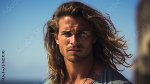 A close-up horizontal portrait of a young handsome man, a surfer with long hair looking at the camera outdoors on a seaside beach. Travel, Vacations, Tourism, Lifestyle concepts.