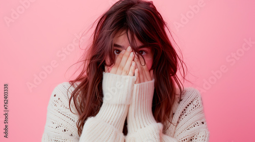 Shy woman peeking through fingers, wearing a white sweater against a pink background