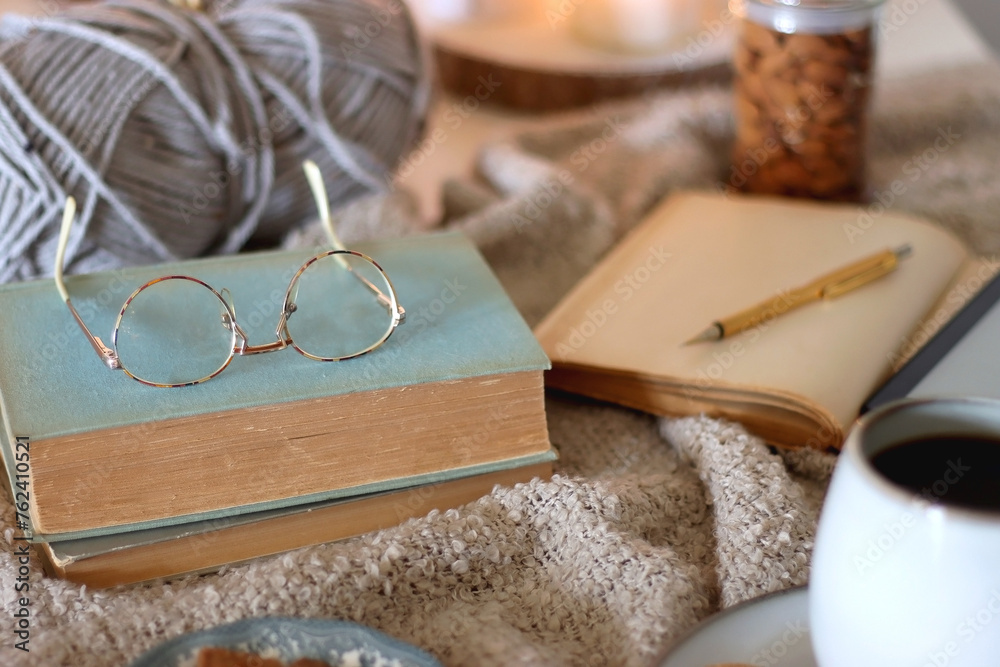 Fototapeta premium Cup of tea or coffee, cookies, books, glasses, e-reader, almonds, ball of yarn, knitting needles and lit candles on the table. Hygge at home. Selective focus.