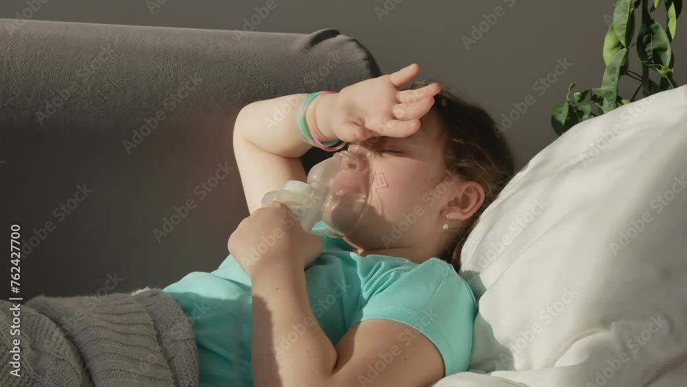 A sick little girl inhales with a nebulizer at home, lying on the couch, exhales medicine through a mask, steam comes out, holds her hand on her forehead, eyes closed A sick little girl inhales with a nebulizer at home, lying on the couch, exhales medicine through a mask, steam comes out, holds her hand on her forehead, eyes closed