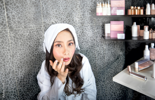 Lot of cosmetics and creams, an Asian woman in a bathrobe gazes at her reflection in the vanity mirror