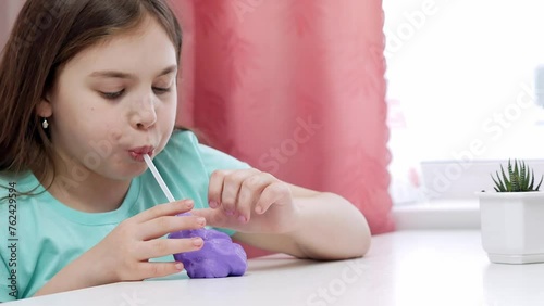 A little girl inflates a large bubble of purple slime through a cocktail tube. A fashionable liquid antistress toy sticks to his hands and fingers.