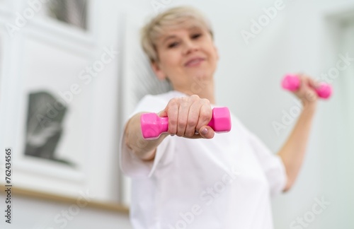 Middle-aged woman in white sportswear exercising with dumbbells at home for health and fitness