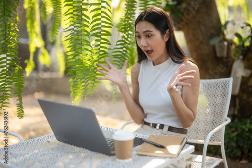 An excited young woman expresses surprise while working on her laptop in a serene outdoor café.