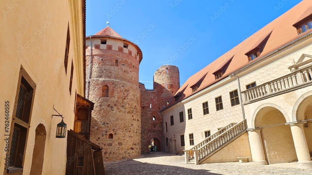 The medieval castle with towers and tiled roofs is built of stone and red brick. The courtyard is paved with stone. The walls have windows and loopholes A staircase with railings leads to the quarters