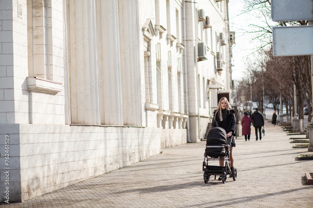 Obraz premium A young mother iswalking around the city with a black stroller and a small child. Woman is carrying a black carriage