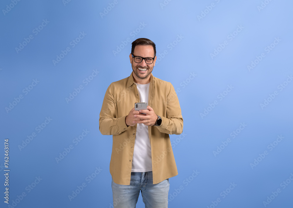 Portrait of smiling young businessman texting online over mobile phone and posing on blue background