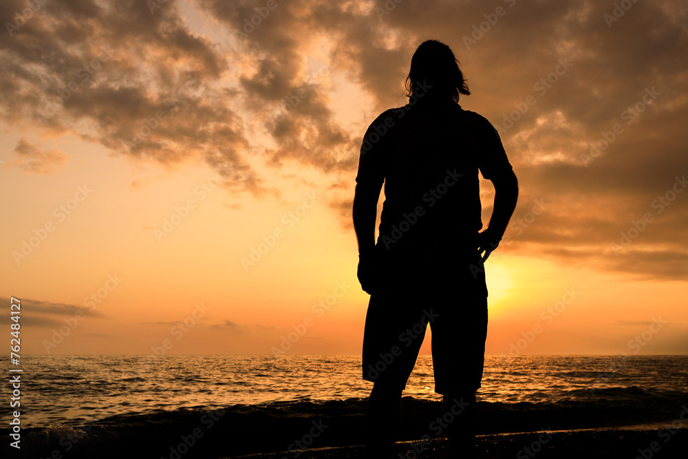 A man stands on a beach at sunset, looking out at the ocean