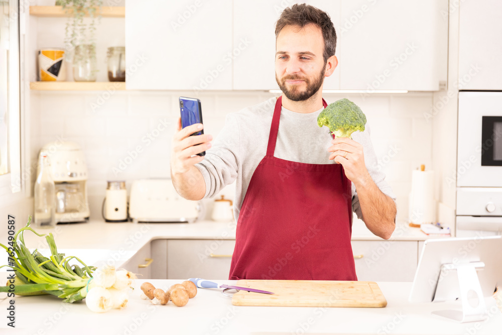 A man is taking a picture of a broccoli in his kitchen. He is wearing a red apron and standing in front of a counter with a knife and a cutting board A man is taking a picture of a broccoli in his kitchen. He is wearing a red apron and standing in front of a counter with a knife and a cutting board