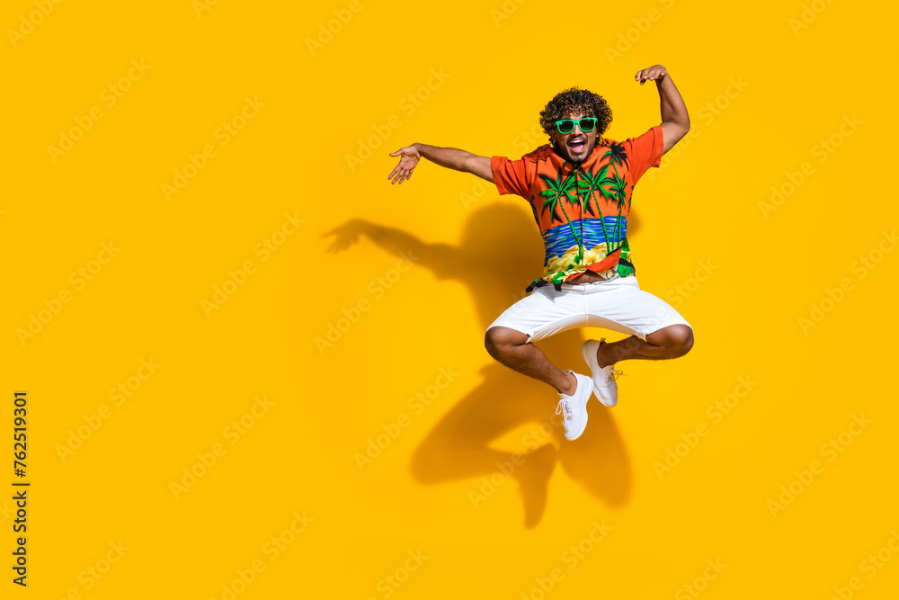 Full length photo of optimistic guy wear hawaii shirt in glasses jumping directing at logo empty space isolated on yellow color background