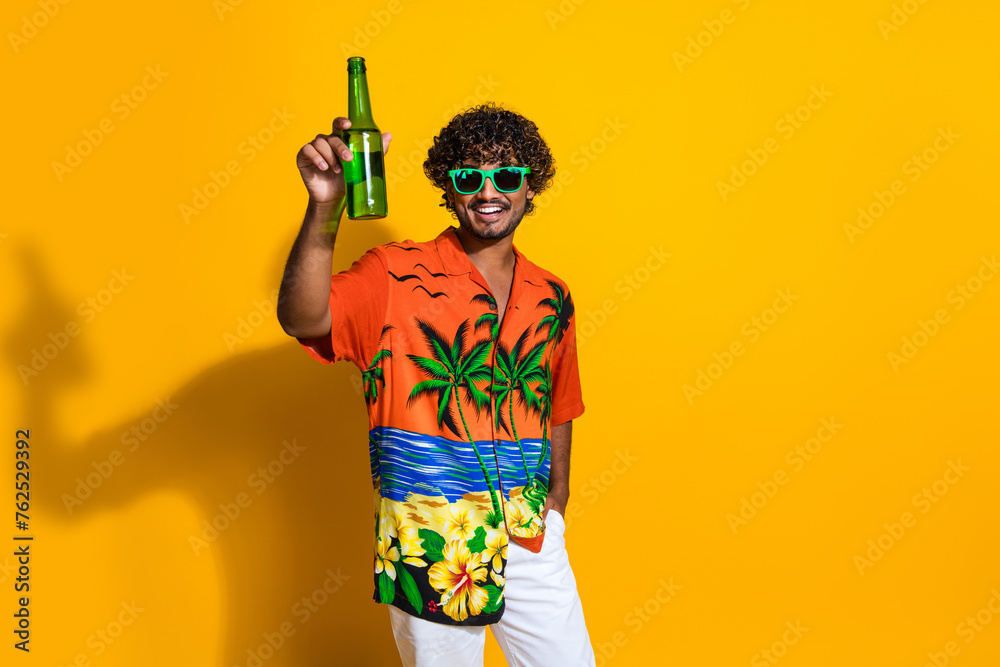 Portrait of handsome guy with afro hair wear hawaii shirt in glasses raising bottle of beer say cheers isolated on yellow color background