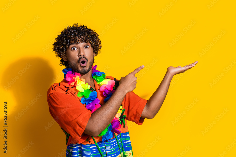 Photo of astonished man dressed print shirt flower necklace directing at offer on arm empty space isolated on yellow color background Photo of astonished man dressed print shirt flower necklace directing at offer on arm empty space isolated on yellow color background