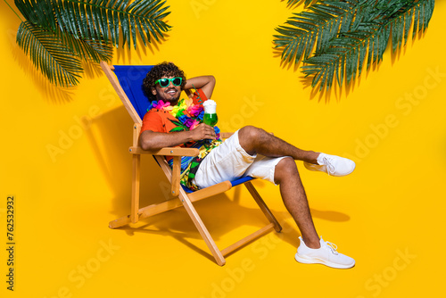 Full size photo of satisfied man wear print hawaii shirt enjoy drink arm behind head lay on deckchair isolated on yellow color background © deagreez