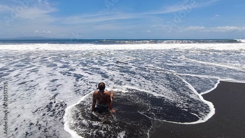 Canvas Print High angle view of a woman sitting alone on a dark black sand beach, surrounded by rushing white ocean foam and crashing sea waves on a sunny day, enjoying tropical coastal nature and vacation