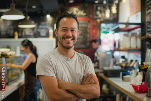 A smiling man with crossed arms standing confidently in a cafe or small business establishment, with an employee working in the background