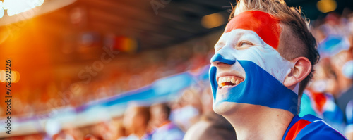 Happy Dutch male supporter with face painted in Dutch flag consists of A horizontal tricolour of red, white, and blue, Dutch male fan at a sports event such as football or rugby match © Pixelmagic