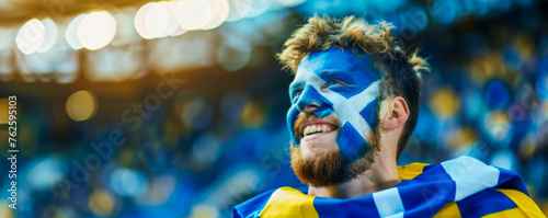 Happy Scottish male supporter with face painted in scotland flag which consists of a white saltire defacing a blue field, Scottish male fan at a sports event such as football or rugby match © Pixelmagic