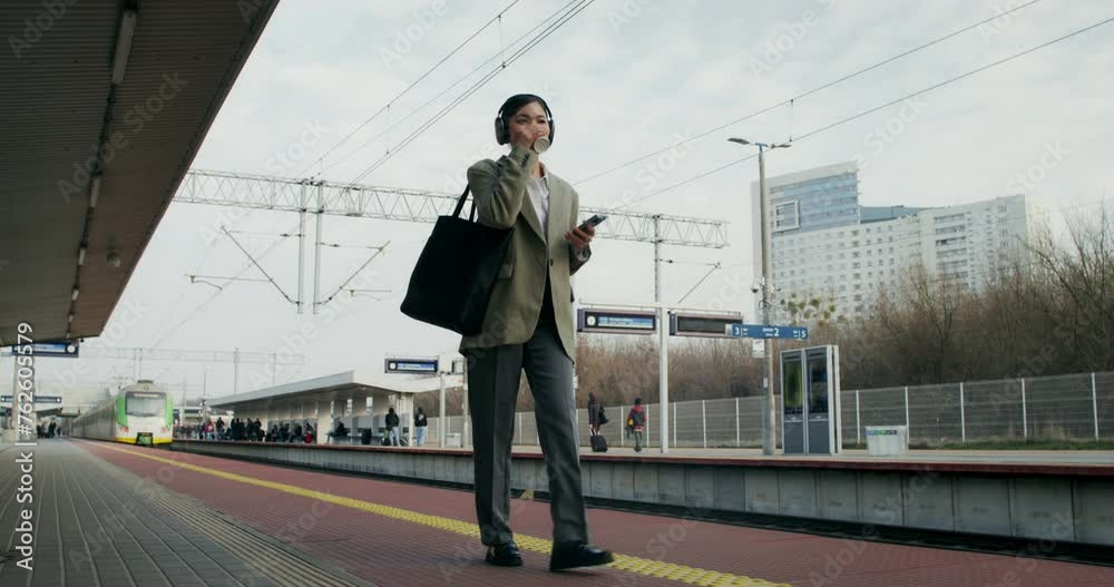 A woman in formal clothes and with headphones on her head uses a mobile phone and drinks coffee from a disposable cup walking along the railway platform