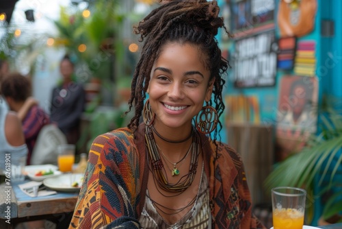 A young woman with dreadlocks and ethnic earrings smiles brightly in a vibrant, bohemian cafe setting.