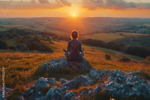 A person meditates on a hilltop at sunset, overlooking a panoramic rural landscape.