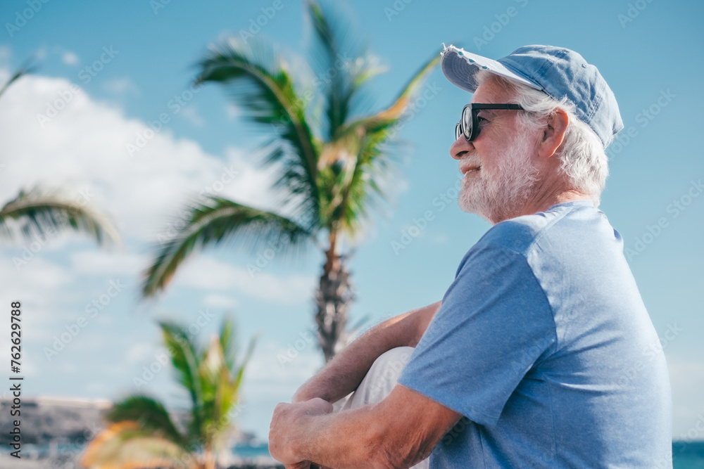 Handsome smiling senior bearded man sitting outdoors face the sea enjoying sunny day, travel, vacation, retirement