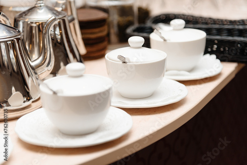 three small white tea cups with lids and spoons on saucers on counter, tableware for cafes, restaurant equipment