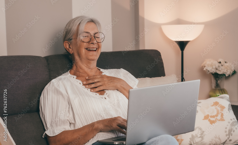 Happy amused senior white haired woman sitting on home sofa watching a funny movie on laptop enjoying tech and social Happy amused senior white haired woman sitting on home sofa watching a funny movie on laptop enjoying tech and social