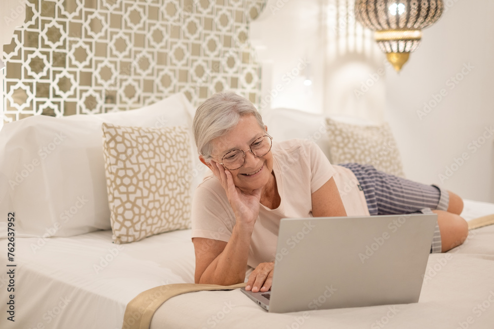 Smiling senior woman lying in bedroom at home or in hotel room browsing internet with laptop enjoying tech and social. Smiling senior woman lying in bedroom at home or in hotel room browsing internet with laptop enjoying tech and social.