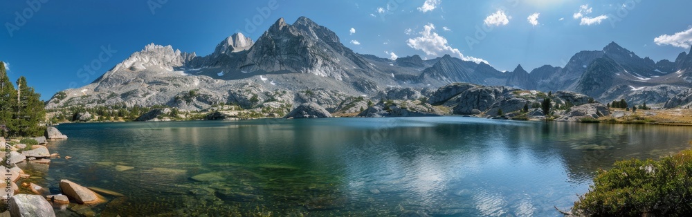 Fototapeta premium A lake is nestled among towering mountains under a clear blue sky. The water reflects the surrounding peaks, creating a stunning natural landscape.