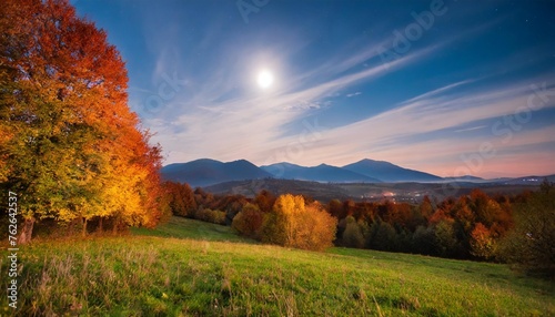 gorgeous countryside at dawn in autumn at night trees in colorful foliage on the grassy field in full moon light mountains in the distance
