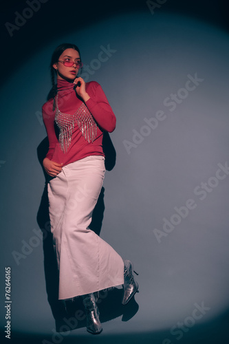 Fashionable confident woman wearing trendy pink sunglasses, turtleneck, rhinestone top, white maxi skirt, silver ankle boots, posing on dark background. Full-length studio fashion portrait