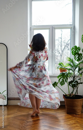 attractive woman with graceful forms in a beautiful dress with a floral print is dancing in a spacious apartment against the background of a bright window