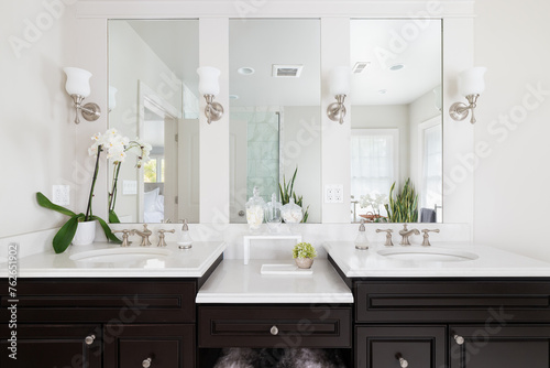 A bathroom with a dark wood vanity  decorations on the white granite countertop  and lights mounted aside the mirror looking at the shower.