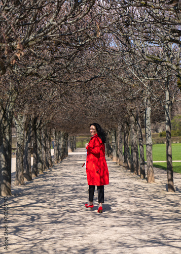 Portrait of a stylish woman outdoors. A model with dark hair, in a red raincoat and red high-heeled shoes walks along an alley among the trees in the park. Turns around and looks at the camera