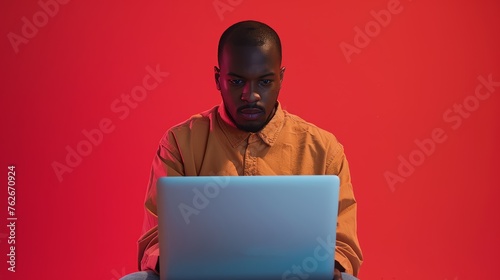 Concentrated man with laptop on red background. Studio shot with bold color scheme. Focused work and digital lifestyle concept.