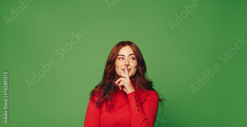 Fashionable woman standing on vibrant green background, quietly gesturing shush with finger