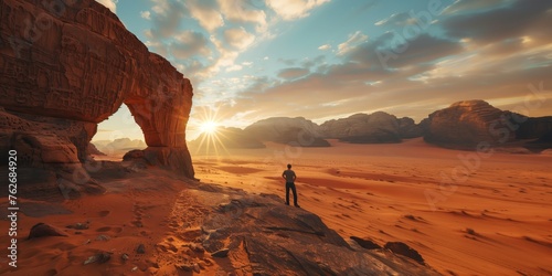 Man standing in the middle of a desert near a rock arch with the sun shining through the arch in the distance, with a mountain in the background. 