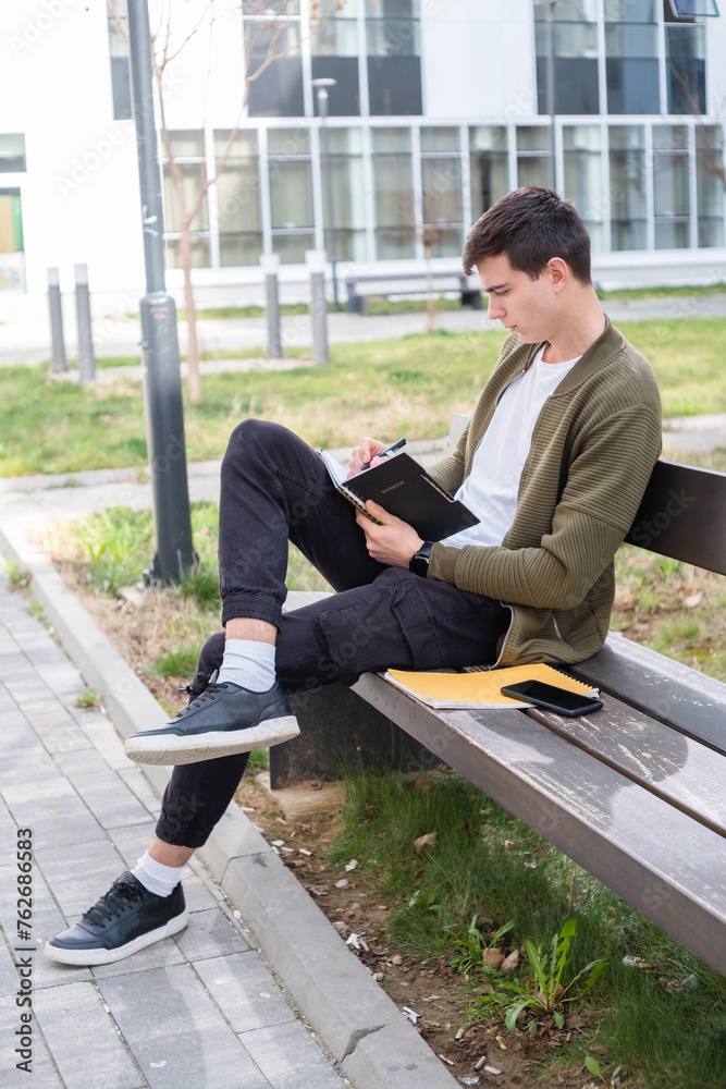 A young male student holding a notebook and sitting on a bench in front of a building, taking notes and learning for the exam on a sunny day A young male student holding a notebook and sitting on a bench in front of a building, taking notes and learning for the exam on a sunny day