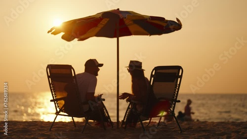 Happy couple on vacation at sea beach. Husband and wife resting, talking at sunny day on ocean shore water, sitting at sunbeds on beach. Family lovers relationships, vacation together, resort concept. photo