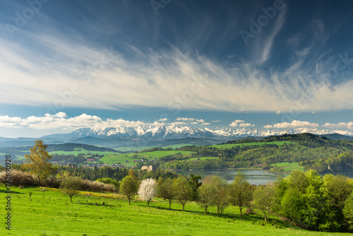 Spring panorama with green fields and trees in Malopolska region, Poland. Czorsztyn castle and snow covered Tatra Mountains behind, Pieniny and beautiful clouds on blue sky.