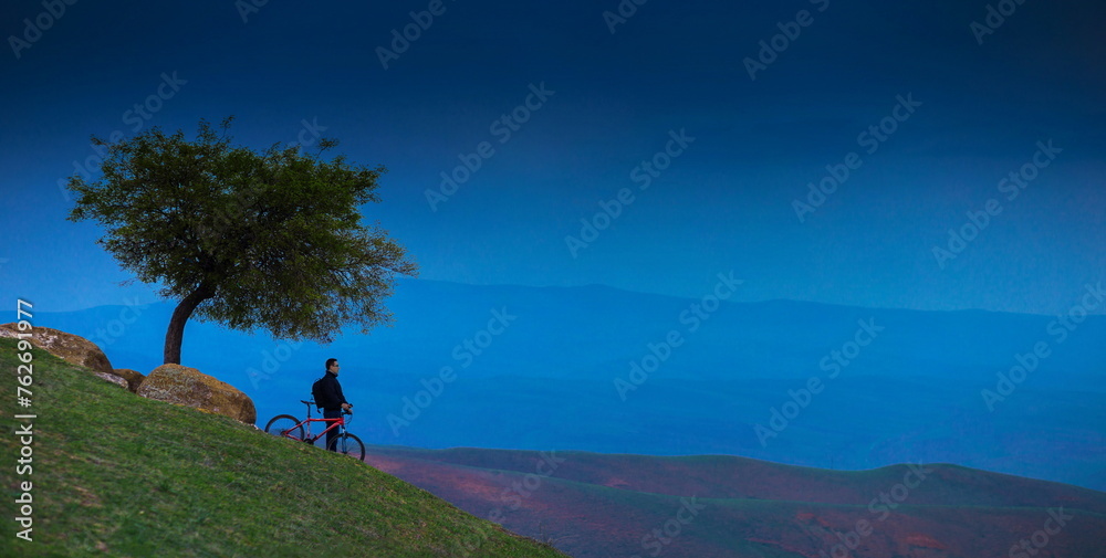 Fototapeta premium A cyclist standing at the edge of a hill near a tree, with a beautiful landscape in the background.