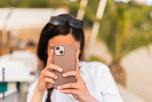 Close-up of a woman in a white shirt using her smartphone, with sunglasses on her head, focused on the screen outdoors.