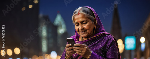 Aged elderly smiling Indian woman with purple tunic using mobile phone on city street at night, blurred out of focus background with bokeh lights, copy space for text, panoramic horizontal banner